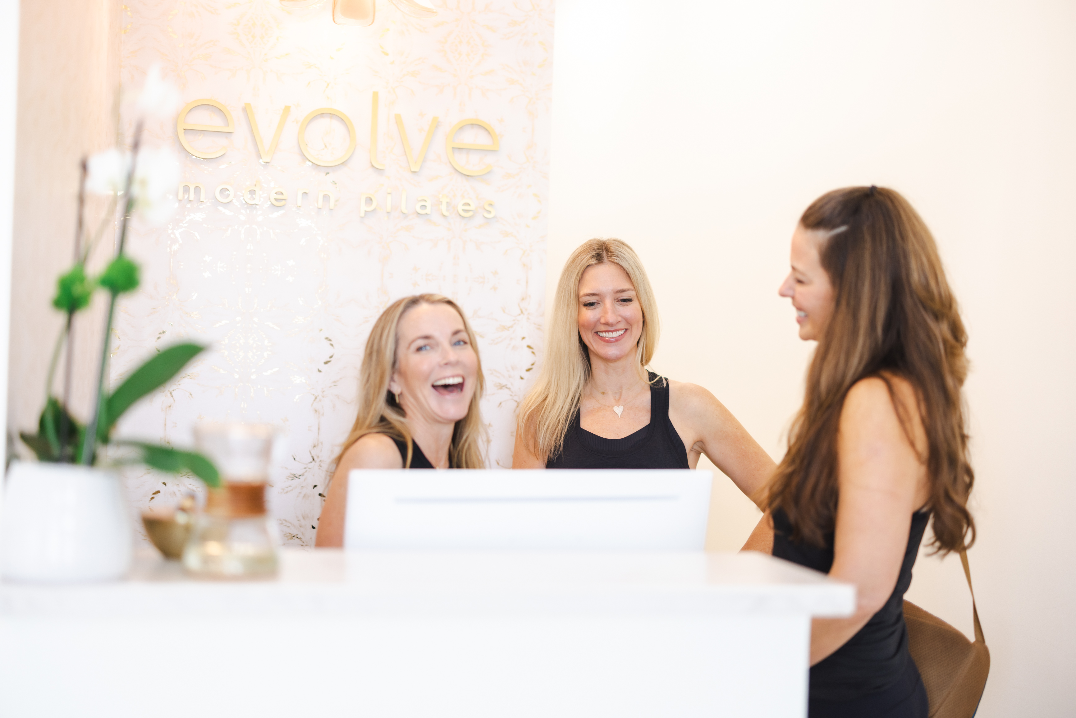 Three staff members smile behind reception desk under Evolve Modern Pilates sign at Charleston studio.