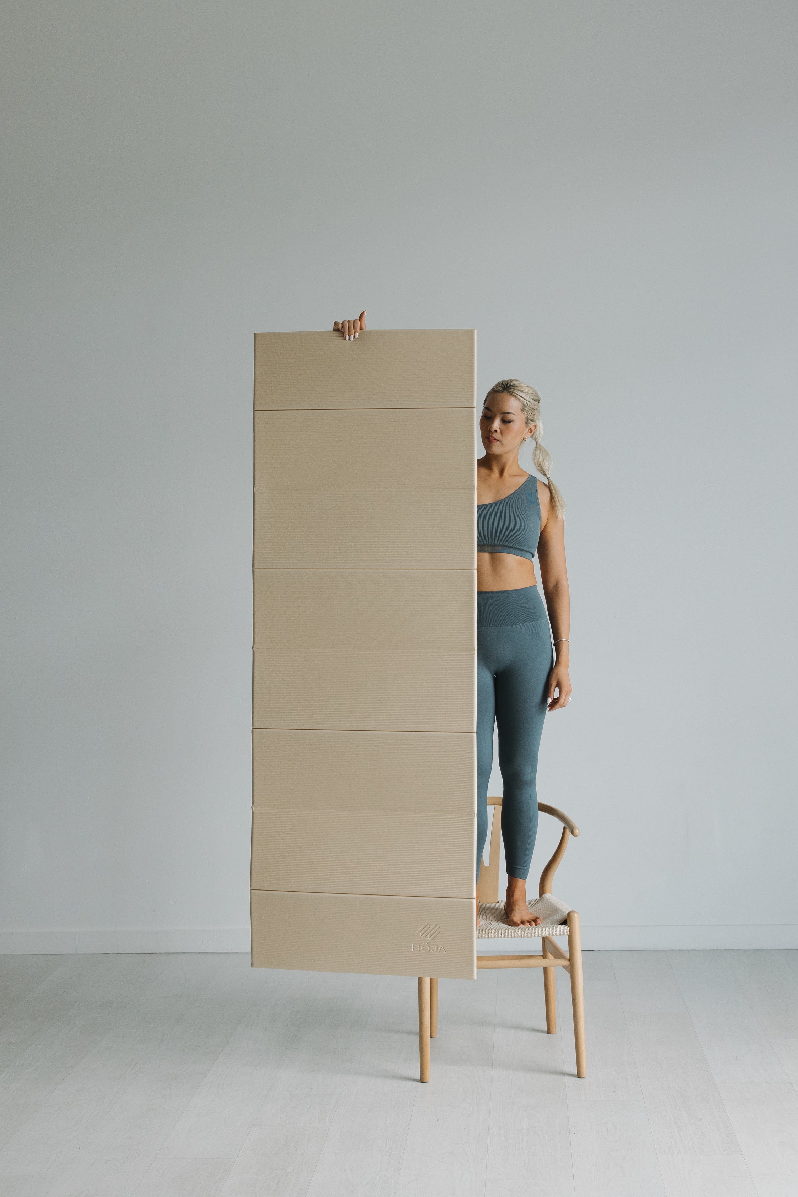 Model on stool next to vertical BOJA yoga mat box illustrating slim, space-saving packaging.