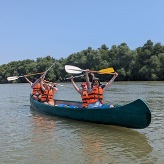 Four REAL School Budapest students in orange life jackets paddle a canoe on a wide river with paddles raised during the REAL Beginnings Adventure, the school's annual rite-of-passage trip held at the start of the year.