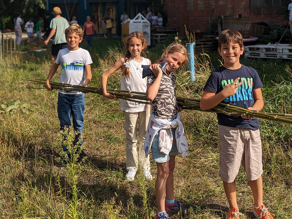 Four REAL School Budapest students smile at the camera carrying long bamboo poles across a grassy outdoor site, during a Dream to Reality project in which students designed and built their own tents before camping overnight.