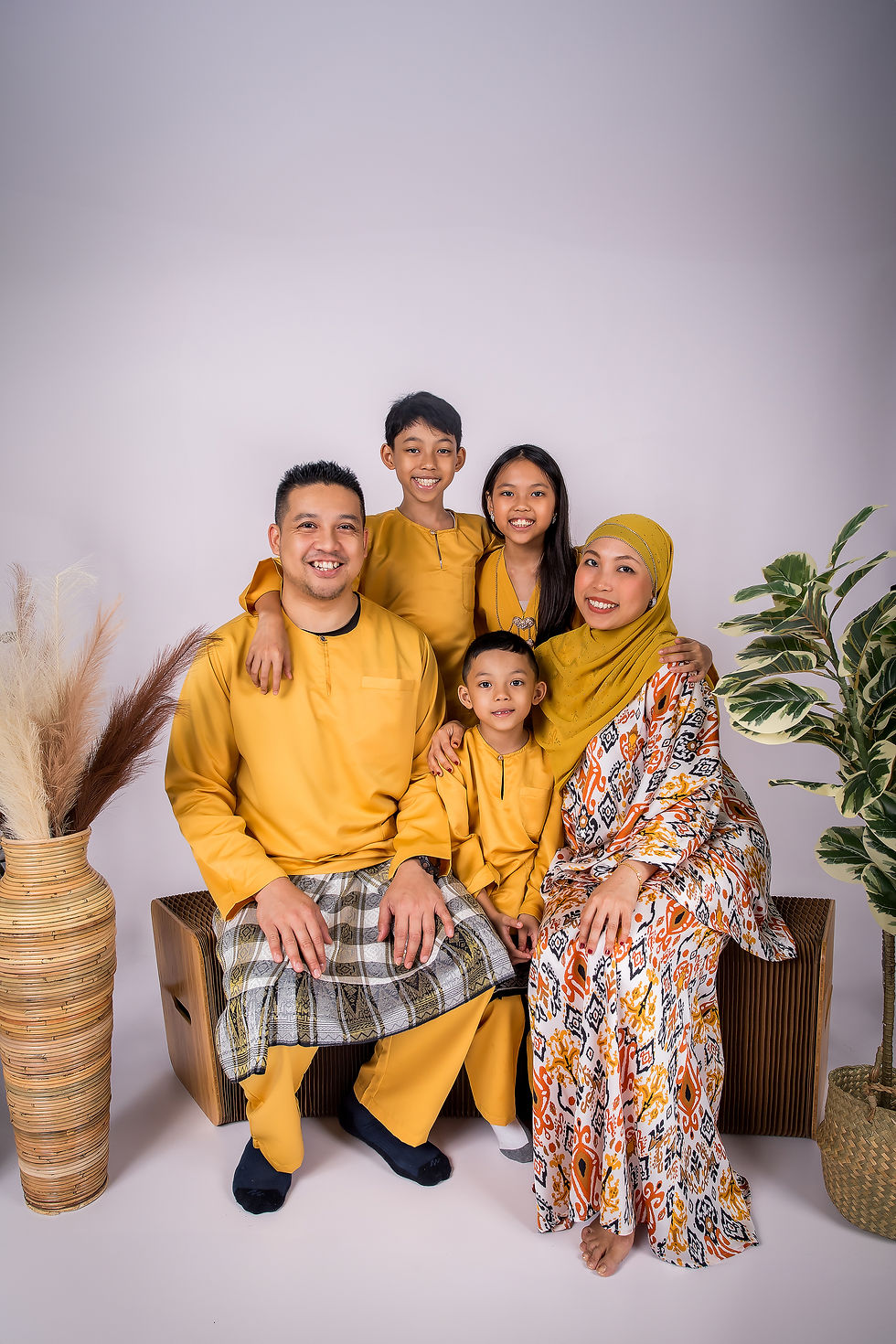 Family of five in matching yellow outfits poses smiling in a studio with potted plants and wicker vases on a neutral backdrop. natural family photoshoot singapore candid interactions