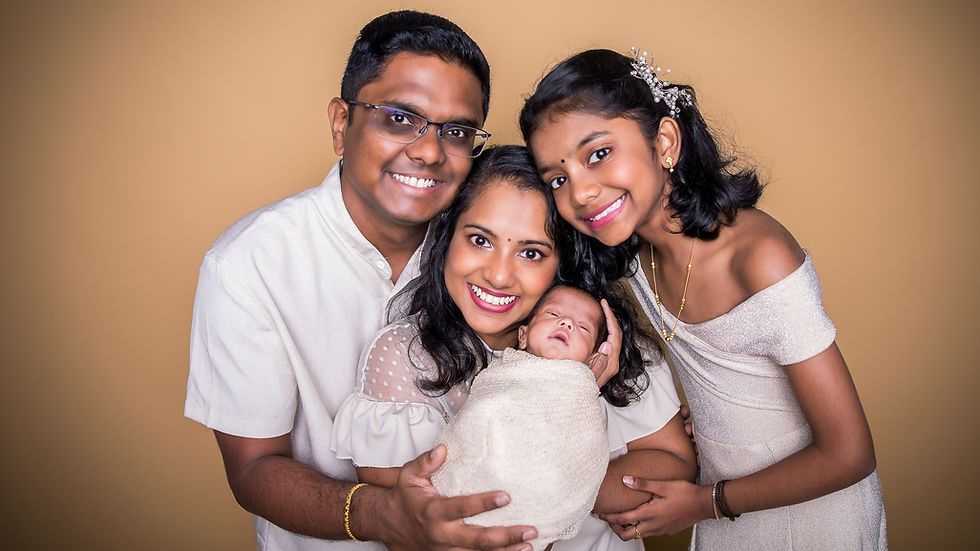 A smiling family of four with a baby wrapped in beige, set against a warm tan background. The mood is joyful and loving. Family newborn photoshoot