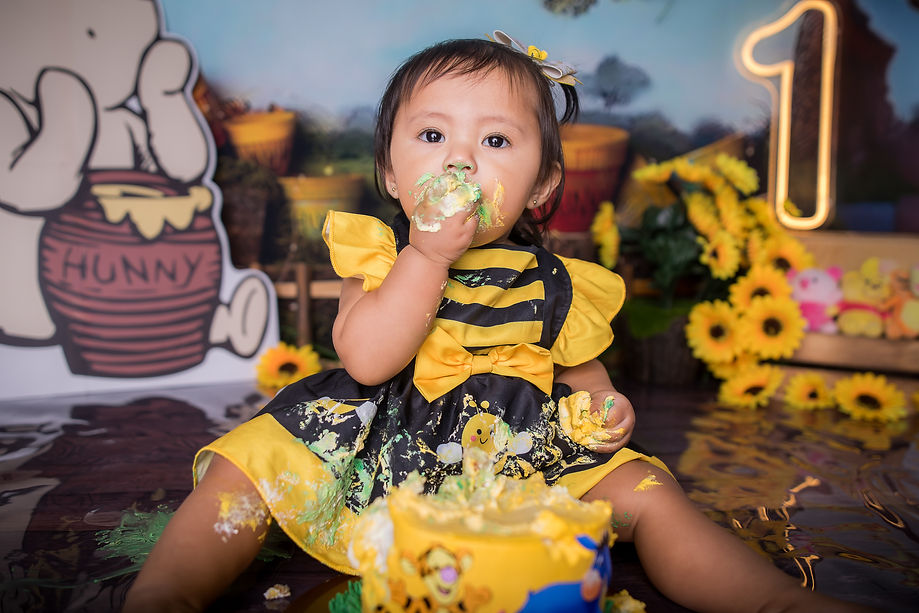 Toddler in bee costume enjoys cake with messy hands. Background features sunflowers, cartoon bear with "Hunny" pot, and large number "1".