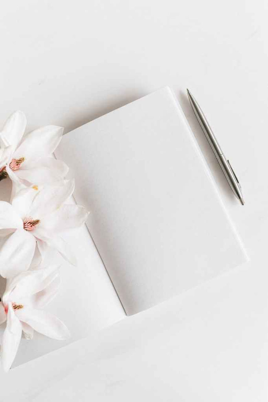 Open notebook with blank pages, silver pen, and white flowers on a white surface. Minimalist and calm aesthetic.