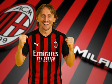 A imagem mostra o jogador de futebol Luka Modric vestido com a camisa do time AC Milan, que tem listras verticais vermelhas e pretas. Ele está sorrindo amplamente e levantando os dois punhos em um gesto de comemoração ou motivação. Ao fundo, há uma grande logo do AC Milan com as letras "ACM 1899" dentro de um círculo, e as listras vermelhas e pretas do time continuam como plano de fundo, com a palavra "MILAN" escrita em branco e levemente desfocada. A imagem transmite uma atmosfera de entusiasmo e apoio ao time AC Milan.