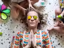 A tired mother lies on the floor with toy blocks on her eyes, hands in a prayer pose, surrounded by playful children and colorful toys—capturing parenting burnout with humor.
