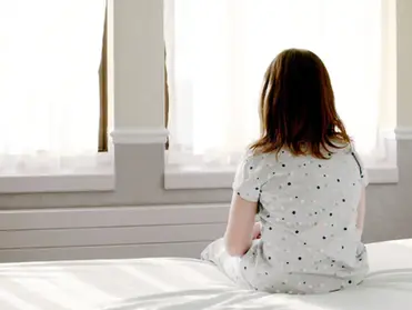 A young girl in polka-dot pajamas sits alone on a bed, facing a bright window with sheer curtains, appearing deep in thought or contemplation.