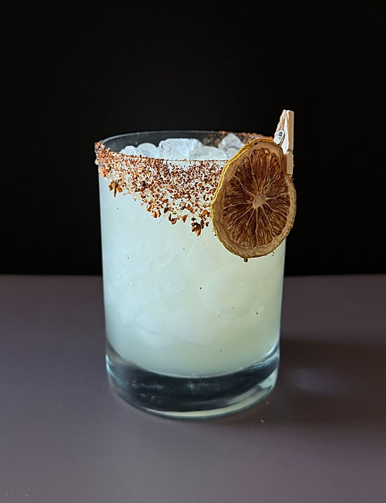 Tommy's Margarita in a stemless rocks glass with a chili salt rim and a fresh lime wedge, filled with ice, against a dark moody black background.