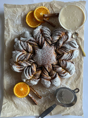 Overhead view of freshly baked cinnamon star bread dusted with powdered sugar on parchment paper, styled with orange slices, cinnamon sticks, a bowl of brown butter glaze, and a fine mesh sifter — cozy holiday presentation on a white surface.