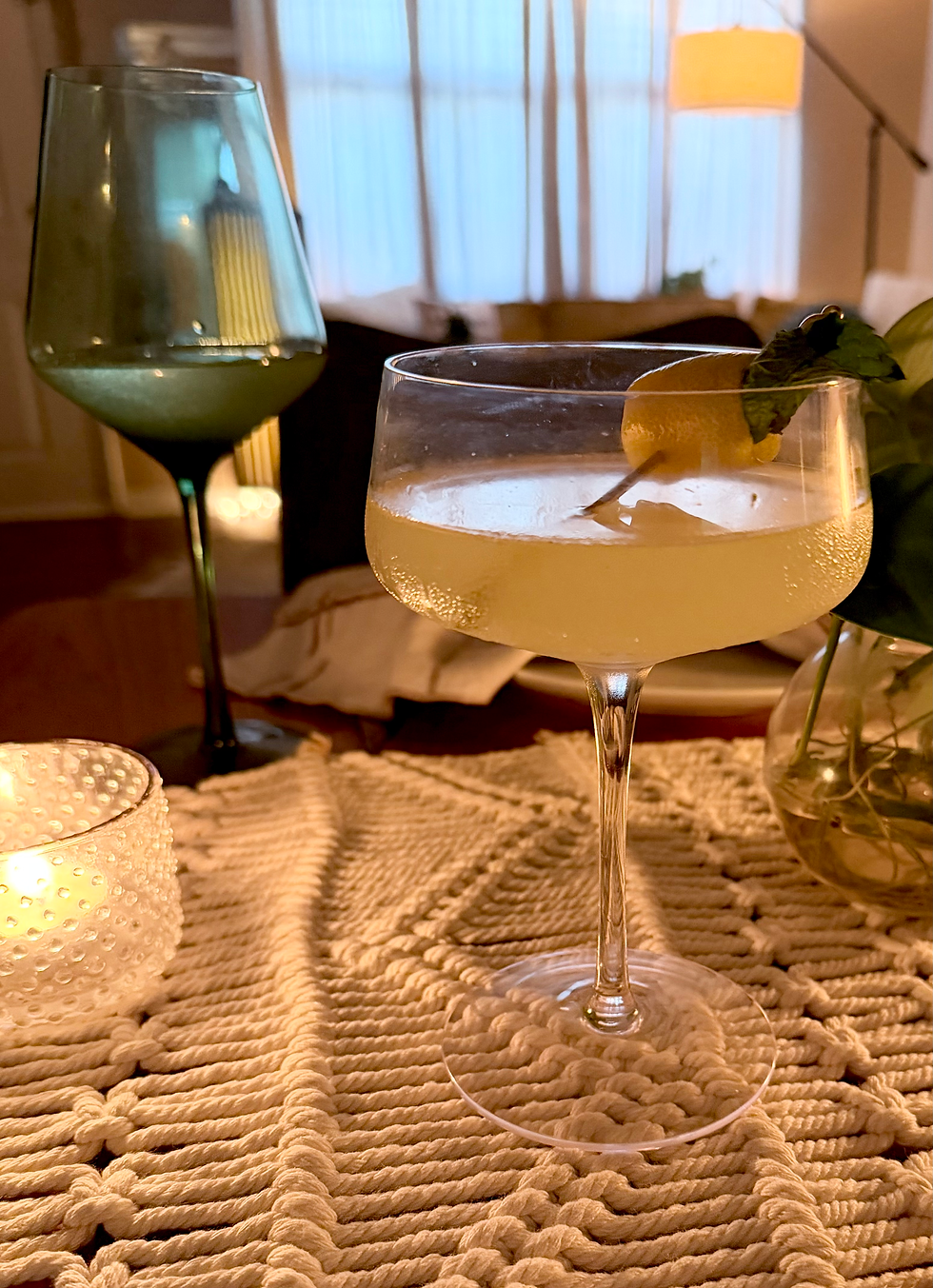 A gin elderflower coupe cocktail with a lemon twist and mint garnish on a candlelit date night table, with a green wine glass and warm lamp glow in the background.