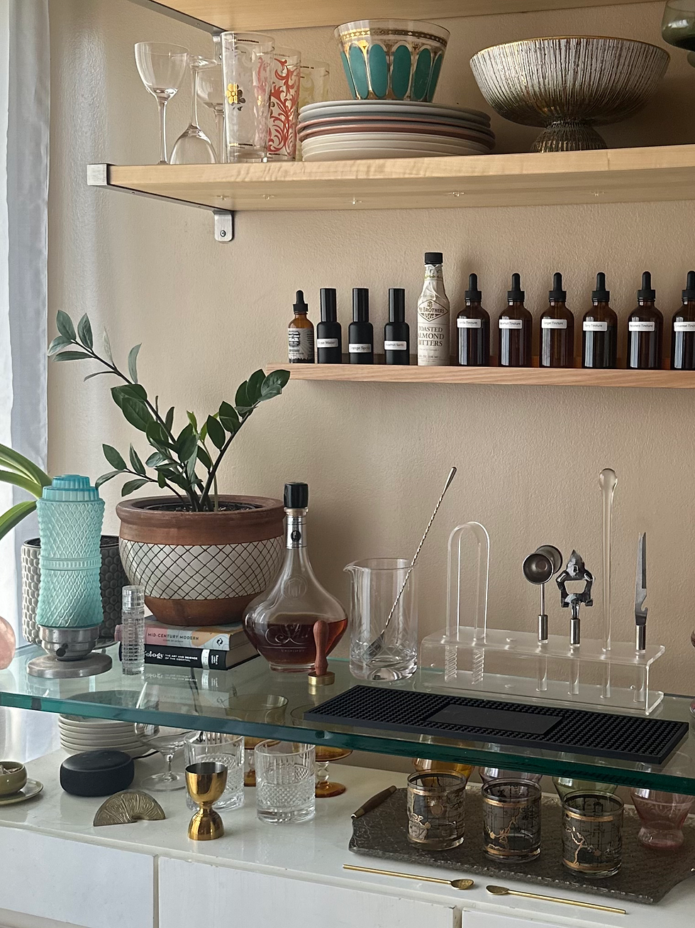Home bar setup with floating shelves, houseplant, cocktail tools, and liquor displayed in a bright kitchen corner