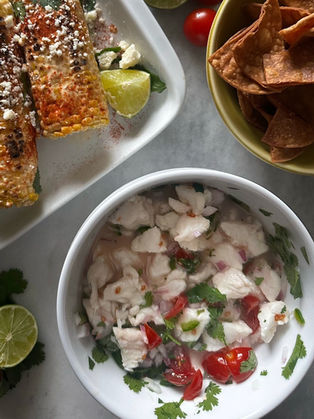 Overhead shot of homemade ceviche in a white bowl with white fish, cherry tomatoes, red onion, jalapeño, and fresh cilantro, styled on a marble surface alongside a green bowl of homemade tortilla chips, elote with cotija and chili, and a halved lime.
