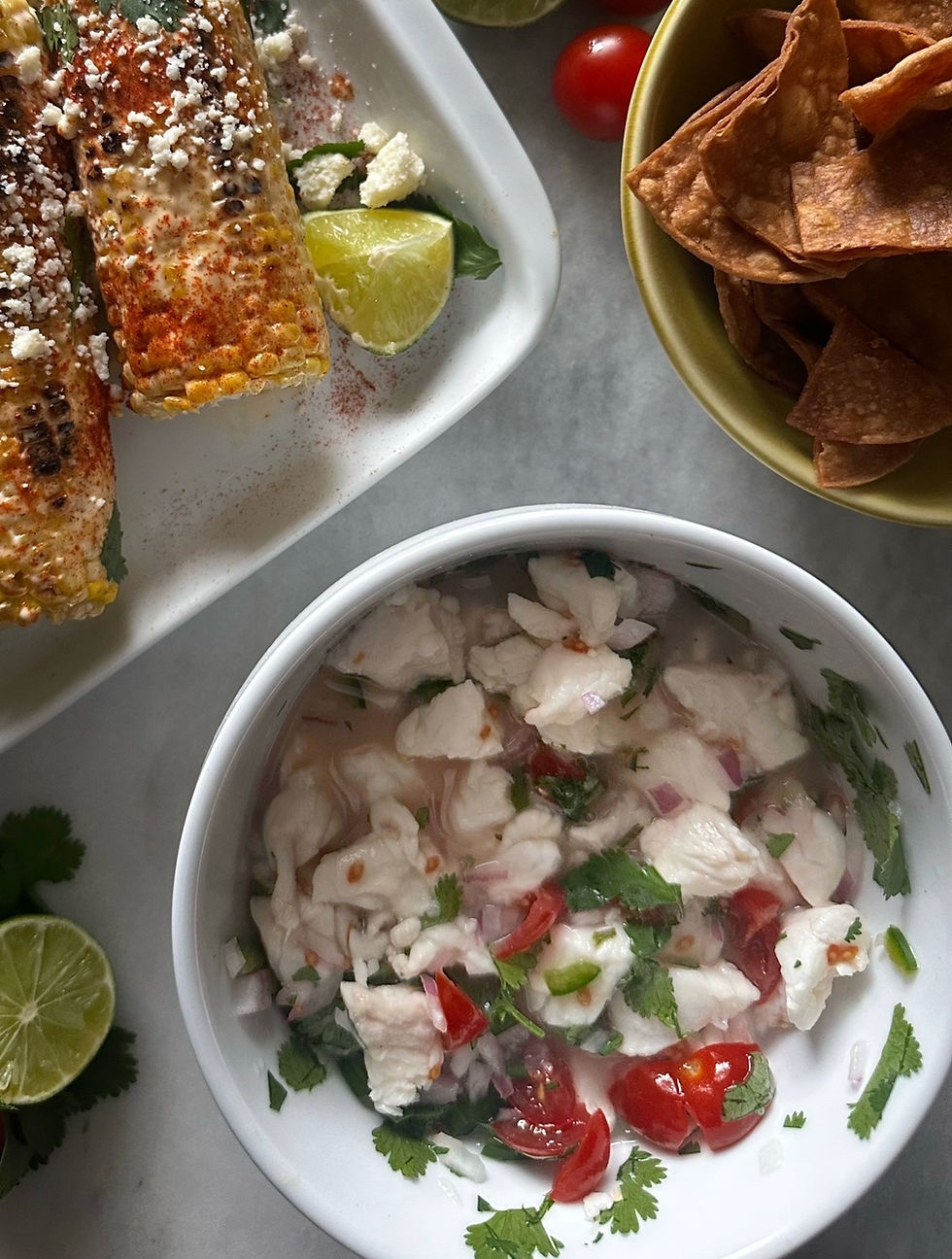 Overhead shot of homemade ceviche in a white bowl with white fish, cherry tomatoes, red onion, jalapeño, and fresh cilantro, styled on a marble surface alongside a green bowl of homemade tortilla chips, elote with cotija and chili, and a halved lime.