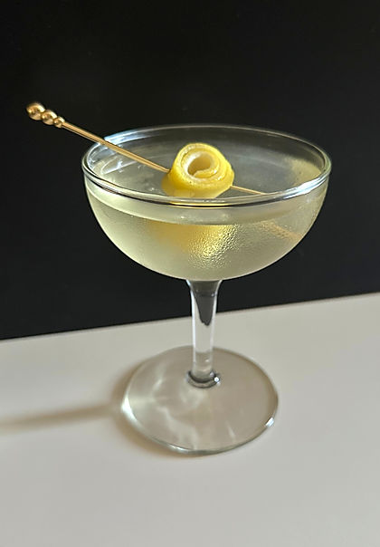 Quit That Racket cocktail in a chilled coupe glass with a pale golden hue, garnished with a coiled lemon twist on a gold cocktail pick, dark background.