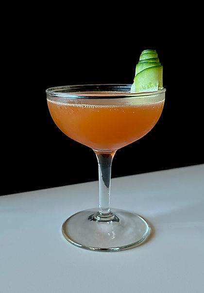 A smoky-orange mezcal cocktail served in a coupe glass with a cucumber ribbon garnish against a dark background.