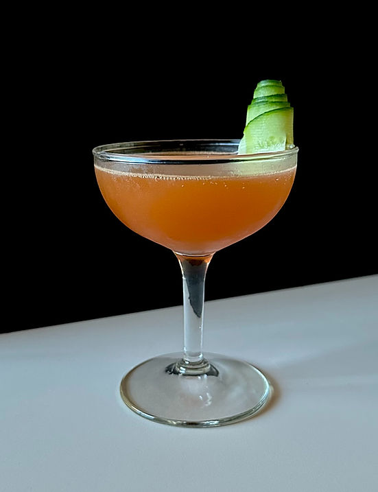 A coupe glass filled with a golden amber mezcal cocktail, garnished with a curled cucumber ribbon against a dark background.