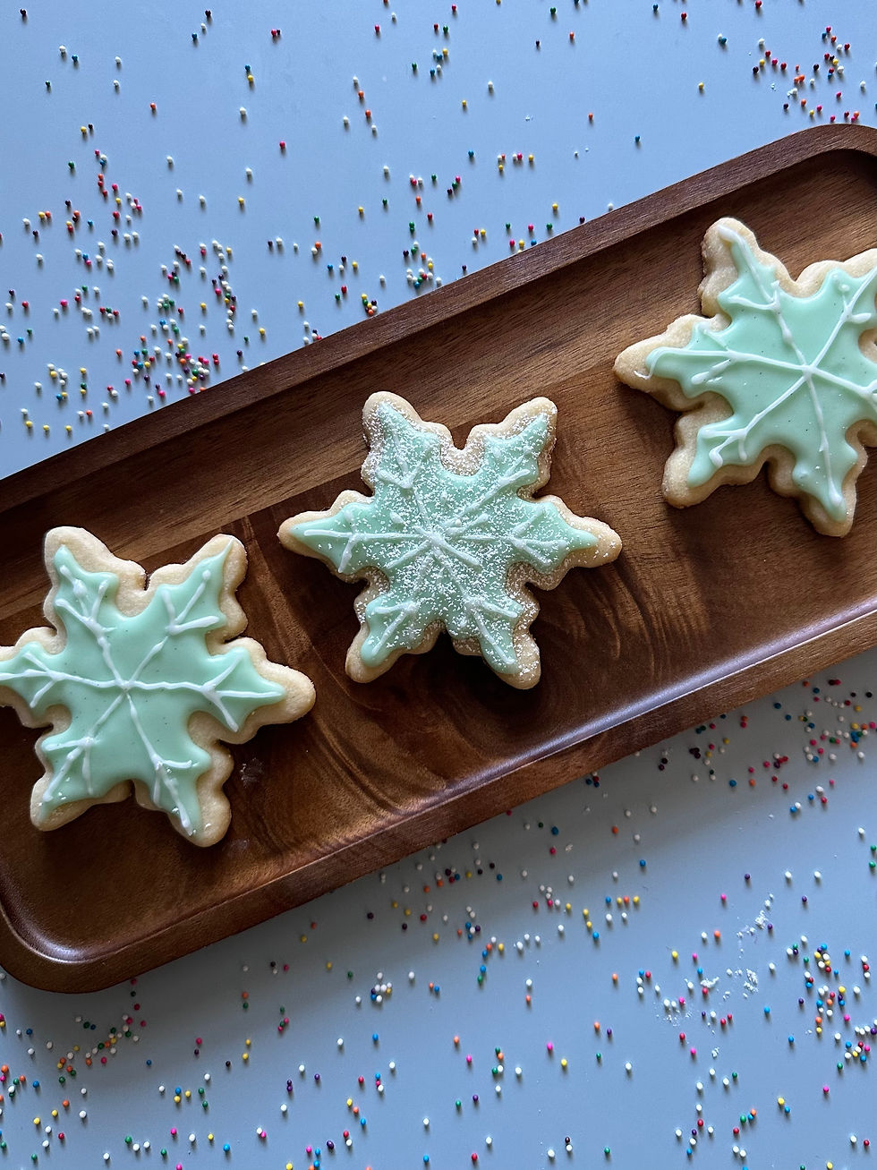 Mint-green snowflake sugar cookies with white icing and sparkling sugar displayed on a wooden tray, surrounded by scattered rainbow sprinkles for a playful holiday touch.