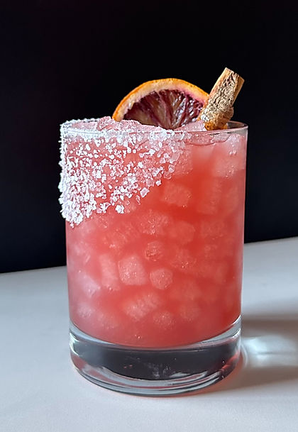 A blood-orange margarita served over crushed ice in a salt-rimmed rocks glass, garnished with a blood orange slice and a cinnamon stick.