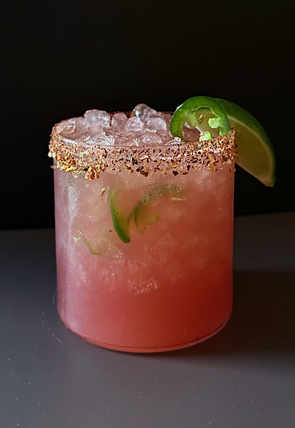 Pink watermelon jalapeño margarita in a rocks glass over crushed ice with a chili salt rim, garnished with a jalapeño slice and lime wedge, dark moody background.