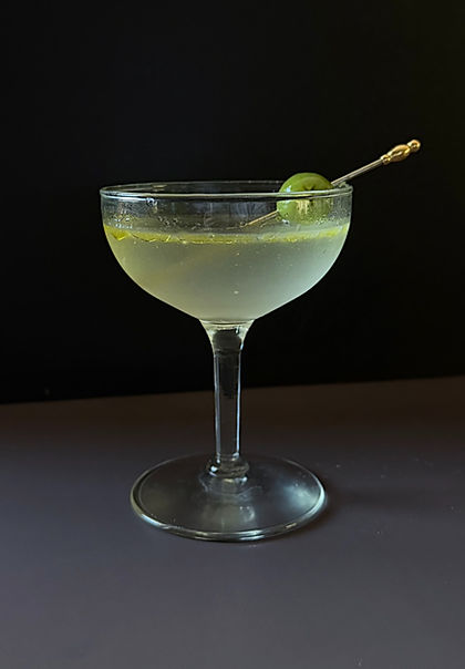 Cucumber gin martini in a vintage coupe glass with a castelvetrano olive on a gold cocktail pick, dark moody background.
