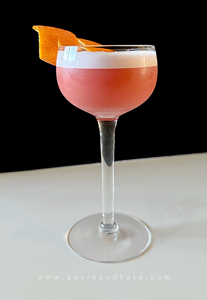 A chilled coupe glass filled with a frothy pink Negroni Sour, topped with a thick white foam layer and garnished with a large curled orange peel, set against a black background.