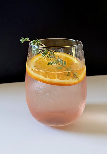 Pale pink Lillet Rosé Spritz in a stemless wine glass over ice, garnished with an orange slice and fresh thyme sprig, dark background with soft white surface.