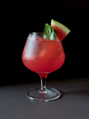 Deep pink watermelon Aperol spritz in a balloon wine glass over ice, garnished with a fresh watermelon wedge and basil leaf, white surface, black background.