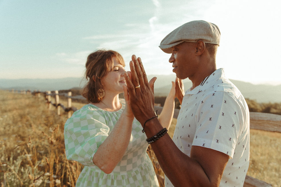Kevin and Kate facing each other with palms pressed together in a golden field at sunset, wedding ring visible, warm light catching both of their faces in a quiet moment of connection and presence.