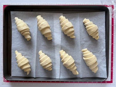 Eight shaped raw croissants arranged on a parchment lined dark baking sheet overhead view, evenly spaced and ready to proof