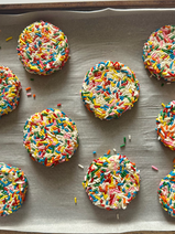 Soft cream cheese cookie dough flattened and covered in colorful sprinkles, arranged on a parchment-lined baking sheet ready for the oven.
