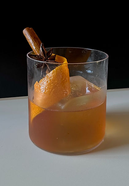 A moody photograph of an apple cider old fashioned cocktail in a clear rocks glass, garnished with a cinnamon stick, star anise, and a large orange peel, set against a minimalist black and beige background.