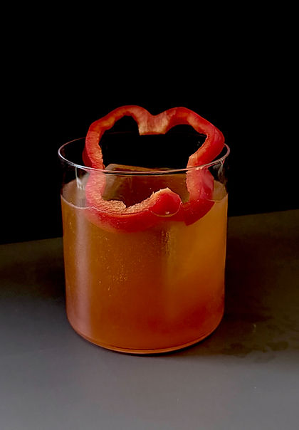 Amber bourbon cocktail in a rocks glass over ice, garnished with a curled red bell pepper slice, dark moody background.
