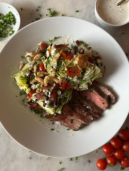 A white dinner plate filled with a wedge salad topped with cherry tomatoes, crispy onions, bacon, chives, and blue cheese crumbles, served alongside thin slices of medium-rare skirt steak. A small bowl of ranch dressing and chopped chives sit nearby on a marble surface.