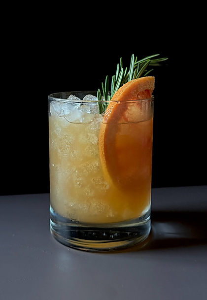 Grapefruit rosemary bourbon sour in a rocks glass over crushed ice, garnished with a half grapefruit wheel and a fresh rosemary sprig, set against a dark moody background.