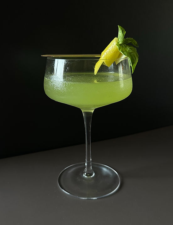 Green herbal cocktail with basil and lemon garnish served in a coupe glass against a dark background