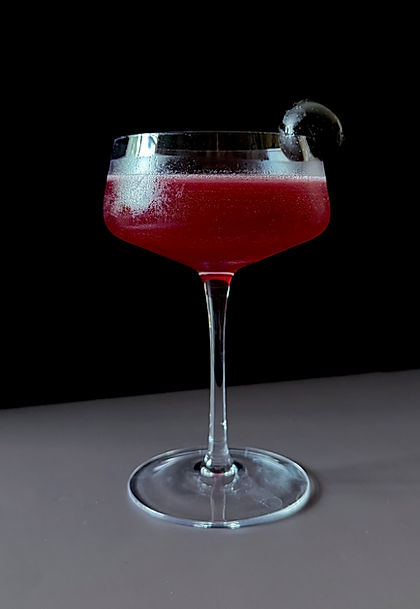 Witch's Currant cocktail in a coupe glass with a black grape garnish, deep red-purple hue against a dark background.