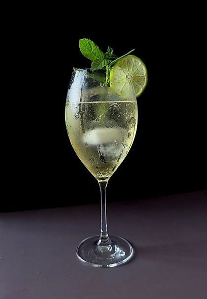 Hugo Spritz in a large wine glass with ice, a lime wheel, and a fresh mint sprig, pale gold and bubbling against a dark moody background.