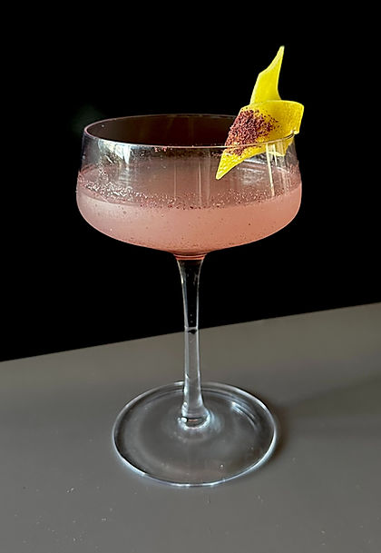 A pale pink sumac gimlet served in a coupe glass, lightly frosted with condensation and garnished with a lemon peel dusted with sumac, set against a dark background.