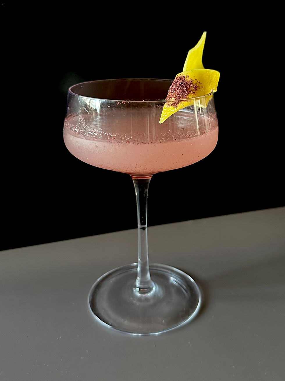Sumac gin sour cocktail served in a coupe glass with a lemon peel garnish dusted in sumac against a dark background.