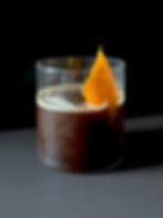 A smoked vanilla espresso old fashioned served in a clear rocks glass over a large ice cube, dark and glossy in color, garnished with an expressed orange peel on a white surface against a dark background.