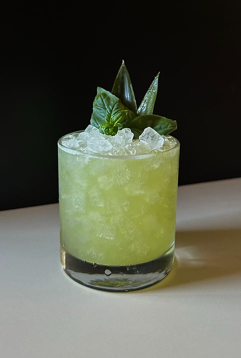 A short rocks glass filled with crushed ice and a bright green pineapple basil cocktail, garnished with basil leaves and a pineapple frond against a black background.