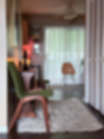 View through a narrow apartment hallway pass-through showing a mid-century drop-leaf table with a glowing lamp and green chair in the foreground, with sheer curtains and a second chair visible in the living room beyond.