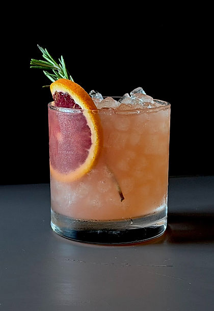 Spiced winter citrus sour in a rocks glass over crushed ice, garnished with a blood orange wheel, a fresh rosemary sprig, and a cinnamon stick, dark moody background.