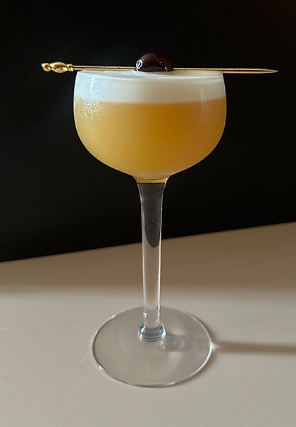 An Amaretto Sour served in a coupe glass with a frothy top, garnished with an Amarena cherry on a gold cocktail pick.