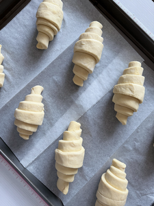 Eight shaped raw croissants on a parchment lined baking sheet from an angled view showing the layered rolls and crescent shapes before proofing