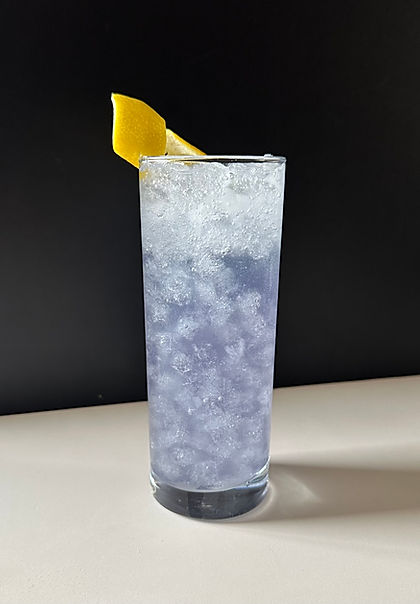 A violet fizz cocktail served in a tall Collins glass filled with crushed ice, soft lavender in color with a frothy top, garnished with a lemon peel against a dark background.