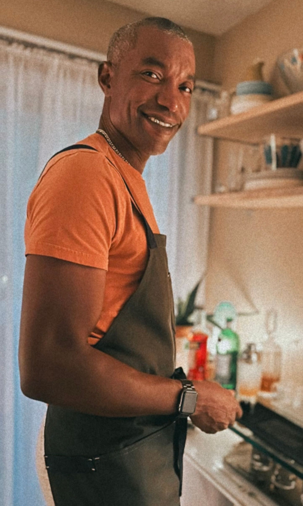 Kevin in an apron smiling back at the camera while cooking in the kitchen, warm natural light, relaxed and at home.