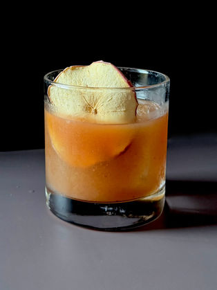 Apple butter old fashioned in a rocks glass over a large clear ice cube, garnished with a dried apple slice, dark background.