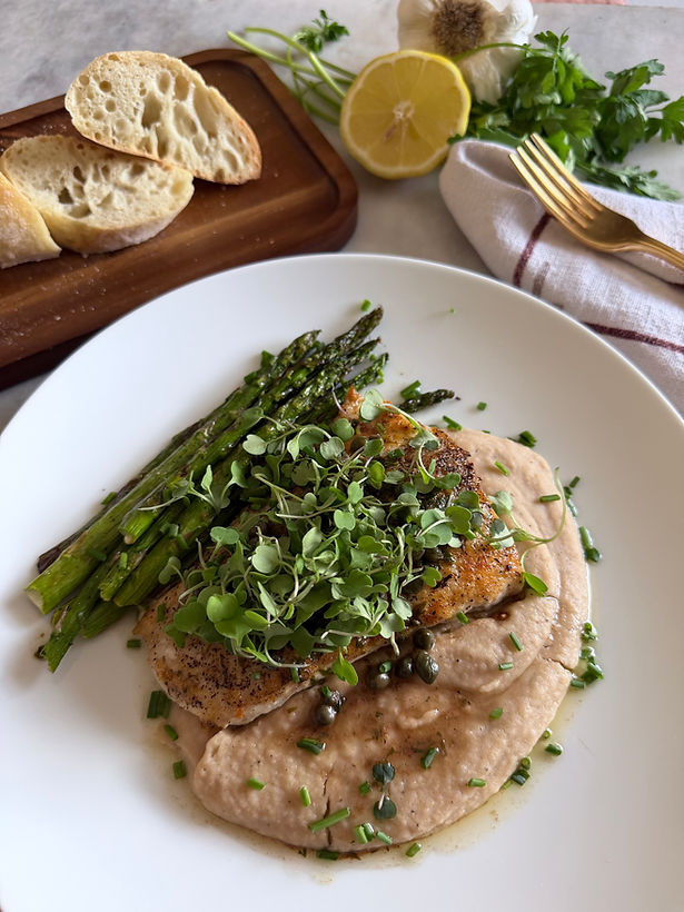 Seared halibut over creamy blended cannellini beans with roasted asparagus, capers, microgreens, and fresh chives in a white bowl, styled with crusty bread, lemon, and a gold fork.