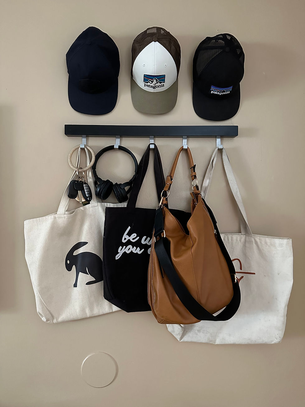 Wall-mounted black hook rail with tote bags, a leather hobo bag, headphones, and keys hanging in an organized entryway, with three baseball caps displayed on the wall above.